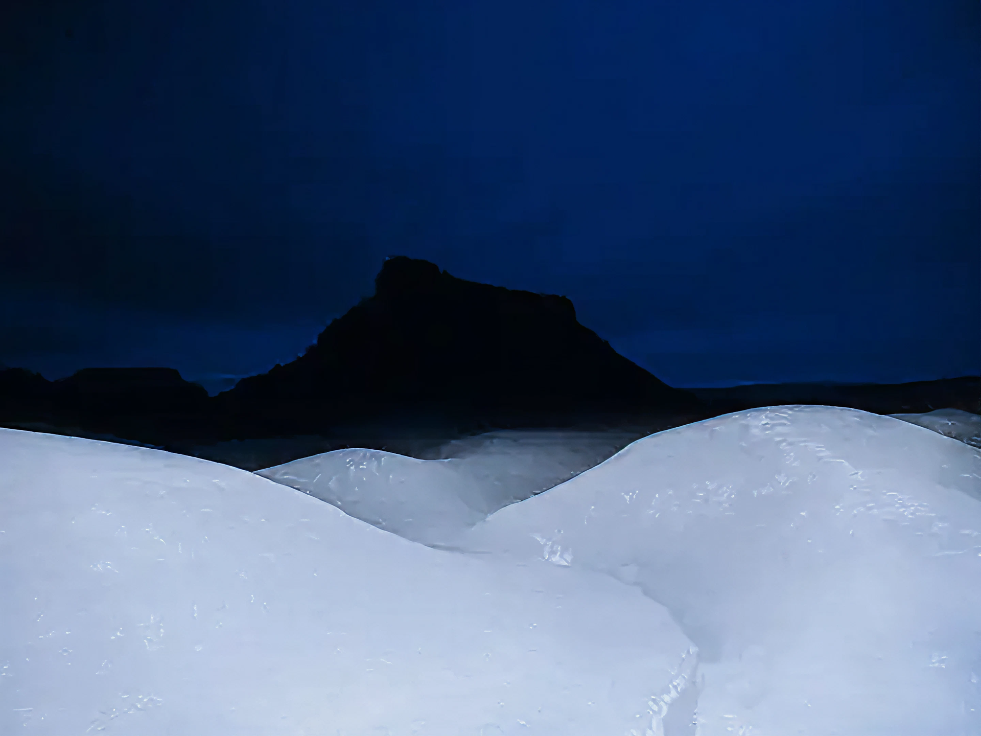 Silhouette of a mountain against a dark blue sky with snow-covered ground.